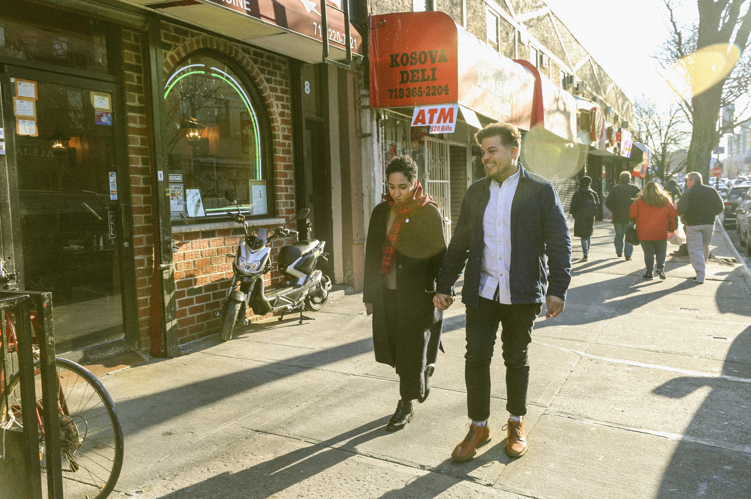 Wide-angle street photograph — couple walking past a Brooklyn deli in low golden sunlight